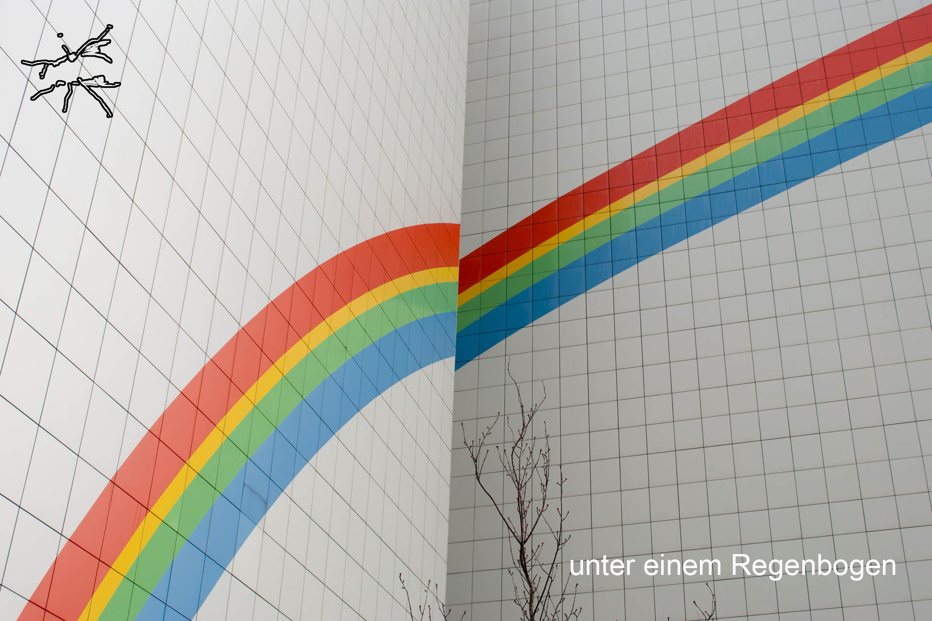 Große Regenbogen-Malerei auf den weißen Kacheln einer Gebäudeecke, darüber kahle Äste im Vordergrund.
