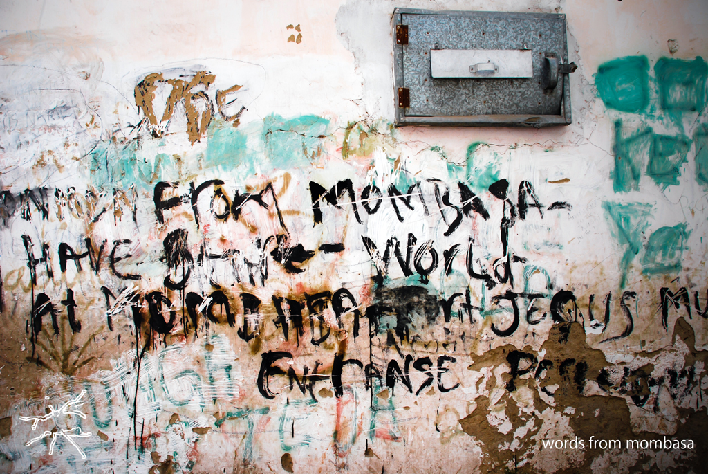 Verwitterte Wand mit farbigen Schichten, kritzelartigen Schriftzügen und gesprayten Worten in Schwarz – Ausdruck, Chaos, Erinnerung.