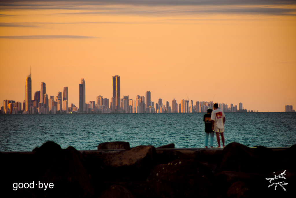 Zwei Menschen stehen eng beieinander auf einer Kaimauer am Meer, den Blick auf eine ferne, von der untergehenden Sonne golden gefärbte Skyline gerichtet. Zwischen ihnen und der Stadt liegt das ruhige Wasser, der Himmel darüber ist zart orange getönt.