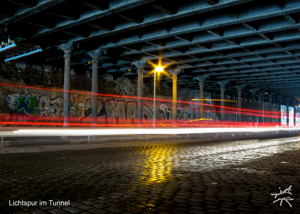 Langzeitbelichtung eines Tunnels mit grauen Stahlträgern, Graffiti an den Wänden und roten sowie weißen Lichtspuren vorbeifahrender Autos.