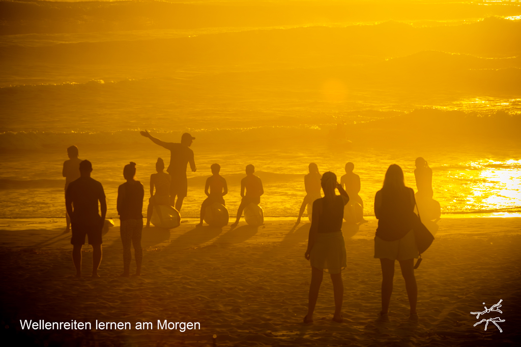 Deutsch:
Am Strand in der Morgensonne zeigt ein Surflehrer mit ausgestrecktem Arm auf das Meer, während junge Schüler aufmerksam auf ihren Boards sitzen. Die Wellen rollen sanft, Eltern stehen im Hintergrund als dunkle Silhouetten.
English:
On the beach in the morning sun, a surf instructor gestures toward the ocean, his arm stretched out, while young students sit on their boards attentively. Waves roll gently, parents stand in the background as dark silhouettes.