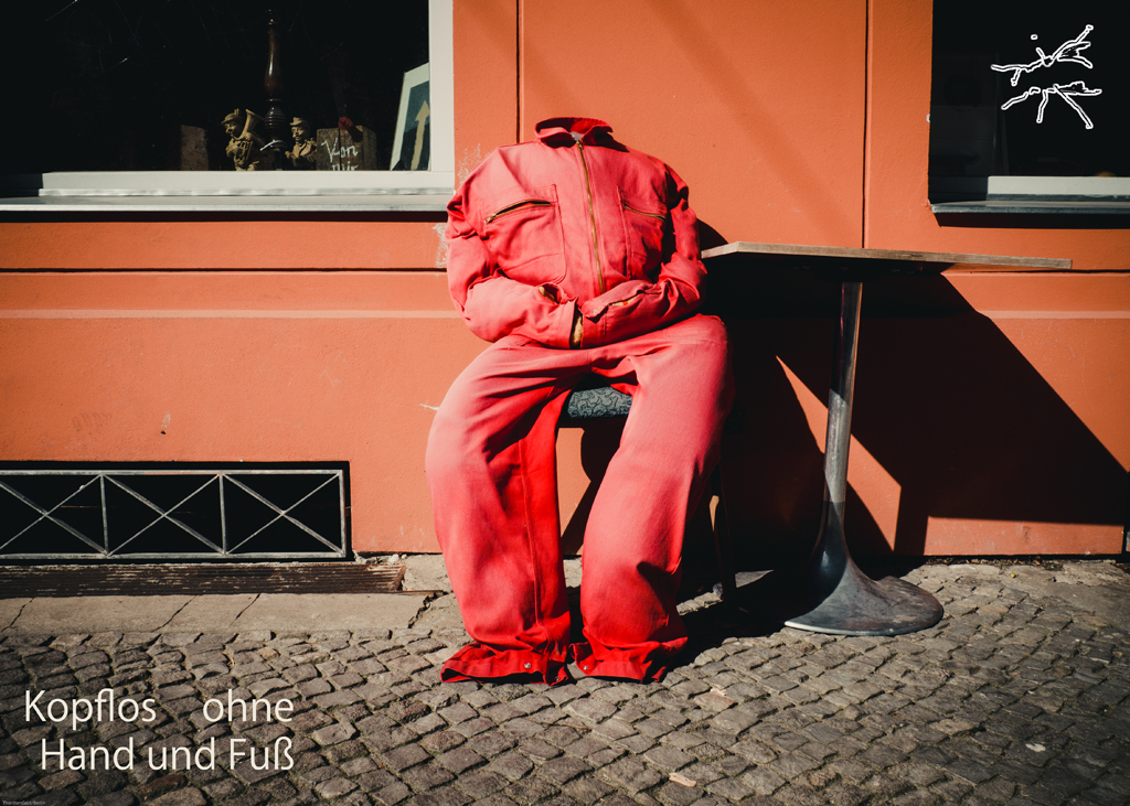 Ein roter Anzug, kopflos und ohne Gliedmaßen, sitzt auf einem Stuhl vor einer orangefarbenen Wand – menschenleer, aber lebendig in seiner Form.