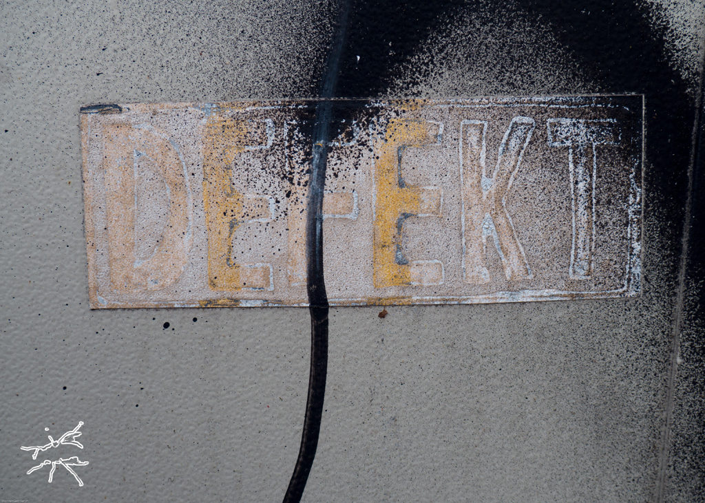 DE: Verblasstes Schild mit der Aufschrift „DEFEKT“, durchzogen von einem dunklen Riss auf grauer Wand. EN: Faded sign reading “DEFECT”, split by a dark crack on a grey wall.