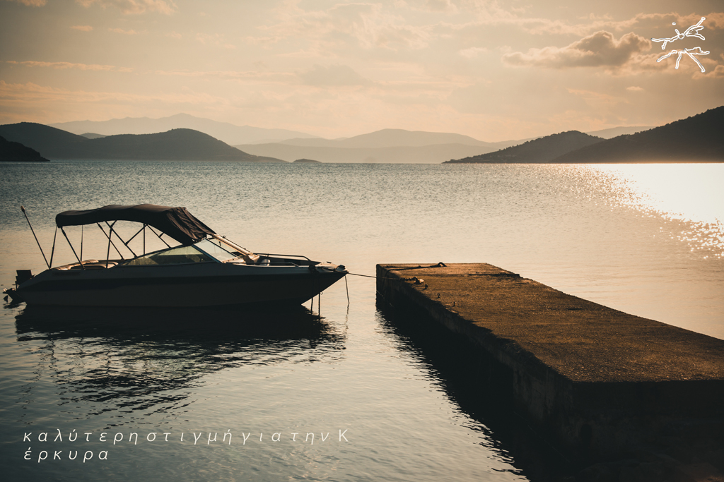 (DE) Ein kleines Motorboot liegt festgebunden an einem schmalen Steg auf ruhigem Wasser, im Hintergrund weite Hügel und sanftes Abendlicht. (EN) A small motorboat is tied to a narrow pier on calm water, with distant hills and soft evening light in the background.
