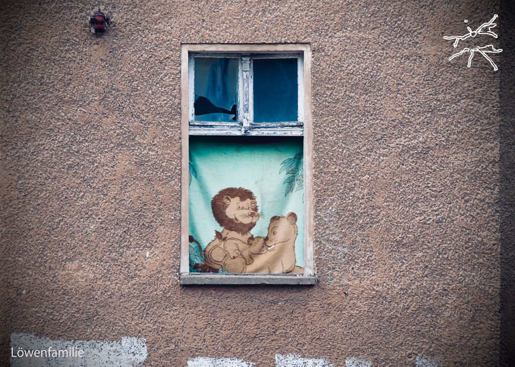 (DE) Ein kleines Fenster in einer alten Hauswand, hinter dem eine Illustration einer Löwenfamilie zu sehen ist. (EN) A small window in an old wall revealing an illustration of a lion family.
