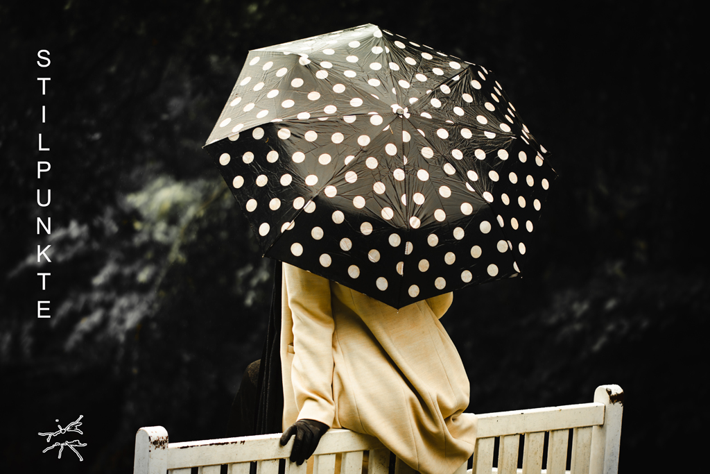 Person mit beigefarbenem Mantel sitzt auf einer weißen Bank und hält einen schwarz-weißen Regenschirm mit Punkten, vor dunklem, unscharfem Hintergrund.