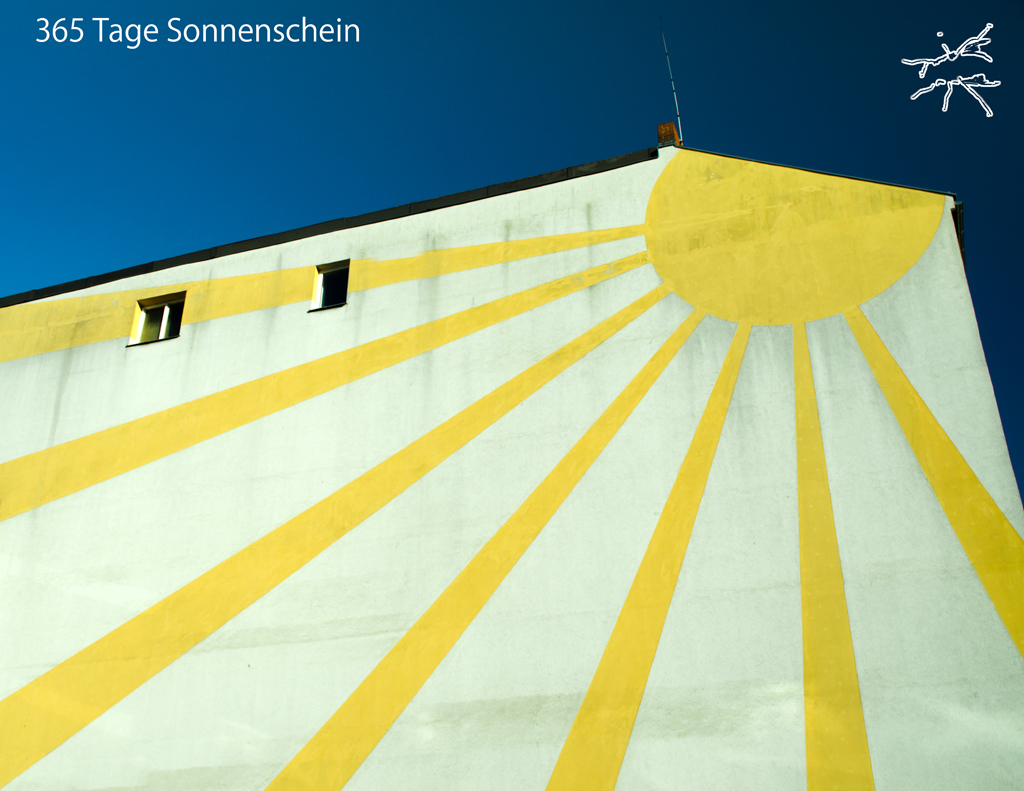 Hausfassade mit gemalter gelber Sonne und Strahlen auf hellem Grund – leuchtend und zugleich nachdenklich im Kontrast zum Himmel.