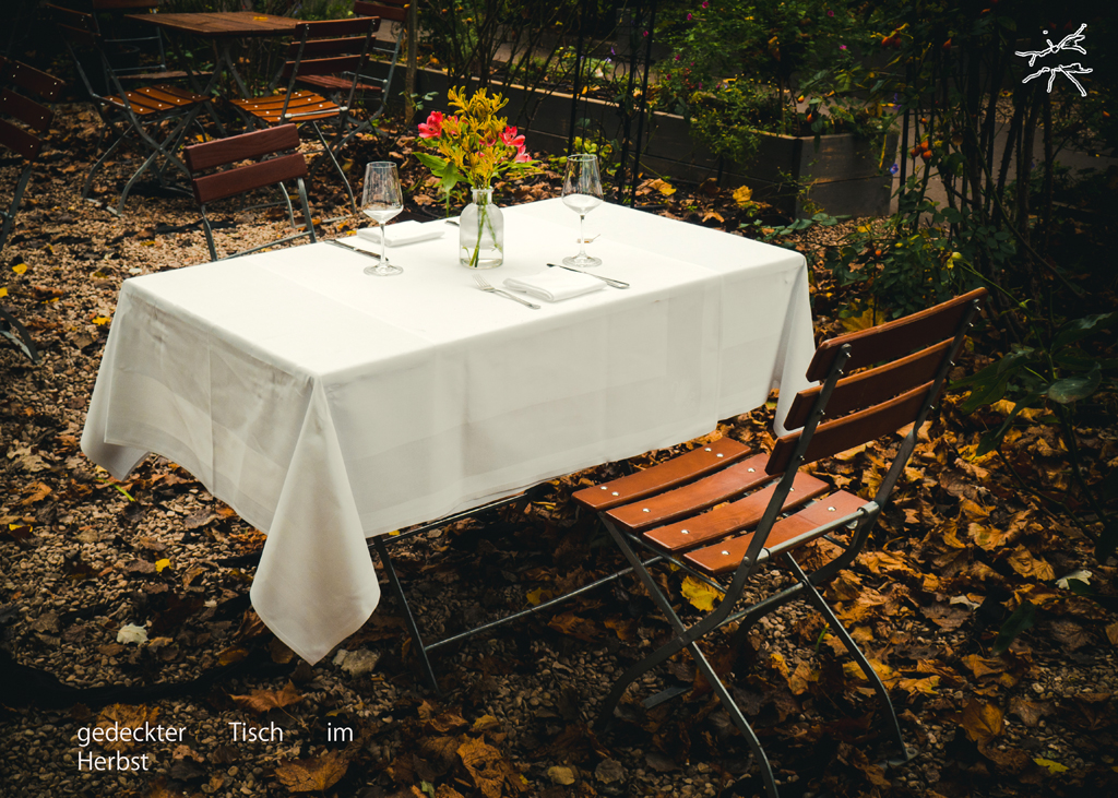 Herbstlich gedeckter Tisch mit weißer Tischdecke im Freien, umgeben von Laub und leeren Stühlen