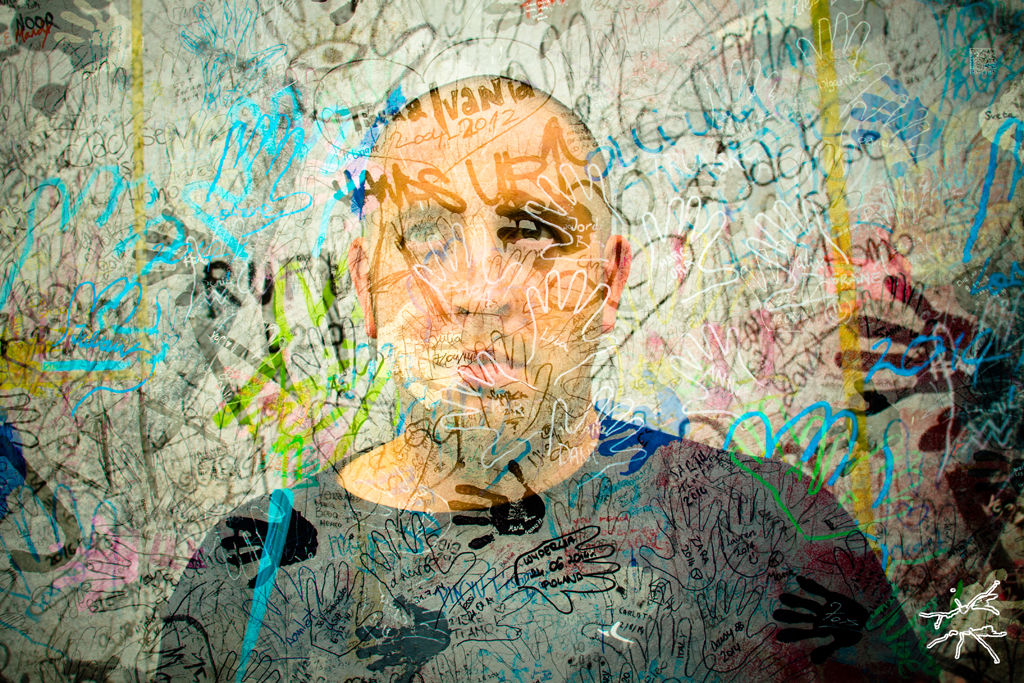 Ein Mann blickt ruhig und ernst durch eine Wand aus bunten Handabdrücken und Graffiti – ein stilles Porträt über Verantwortung, Spuren und kollektive Energie.