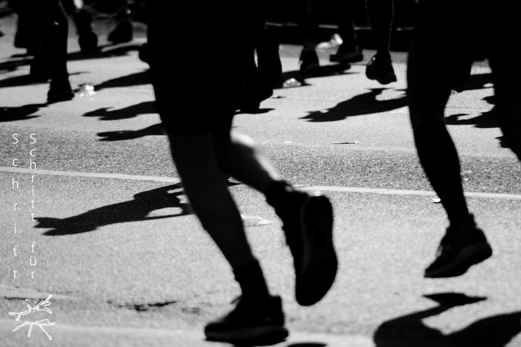 Schwarz-weiß Fotografie von laufenden Menschen, deren Schatten auf die Straße fallen. Die Bewegung und Dynamik ist durch die Unschärfe der Beine spürbar.