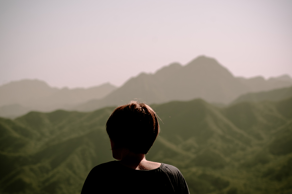 Eine Person steht mit dem Rücken zur Kamera, blickt über eine weite, grüne Hügellandschaft. Das Licht ist weich, die Atmosphäre ruhig – als wäre dies ein Moment des Innehaltens. Der Text überlagert das Bild, spricht von Begegnungen, ungesagten Worten und flüchtigen Augenblicken, die in ihrer Echtheit bleiben.