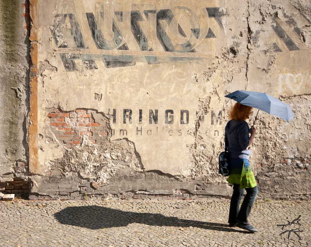 Deutsch: Eine Frau mit rotem Haar und blauem Regenschirm geht an einer verwitterten Hauswand vorbei. Der Putz blättert ab, alte Werbeschriftzüge leuchten durch. Sonne fällt schräg auf die Szene, wirft einen langen Schatten auf das Pflaster. English: A woman with red hair and a blue umbrella walks past a weathered wall. The plaster is crumbling, faded advertisement letters emerge. The sun hits the scene at an angle, casting a long shadow on the cobblestones.