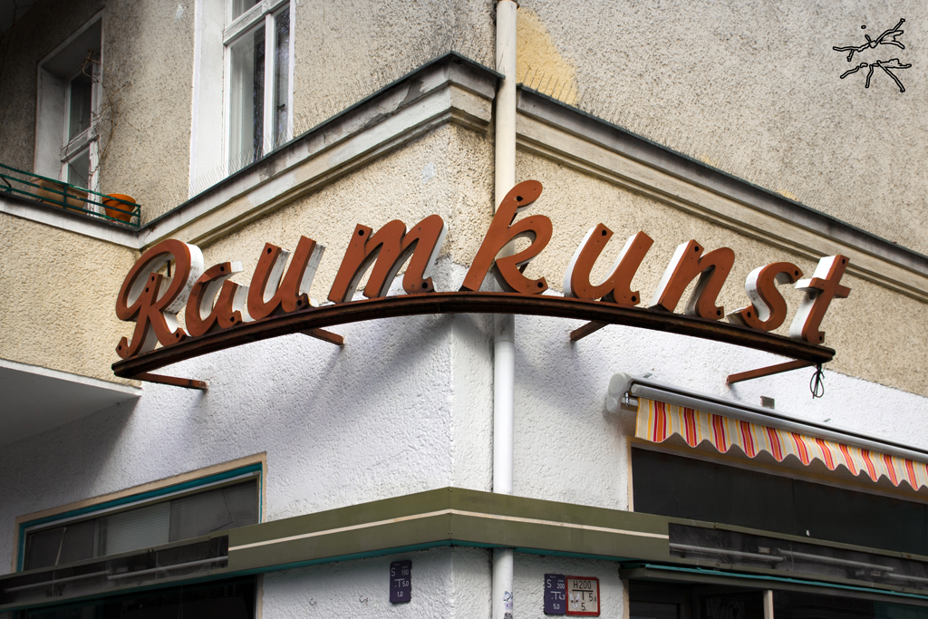 Altes Ladenschild mit Schriftzug „Raumkunst“ an der Hausecke, rostrote Lettern auf gebogener Halterung – nostalgisch und urban zugleich.