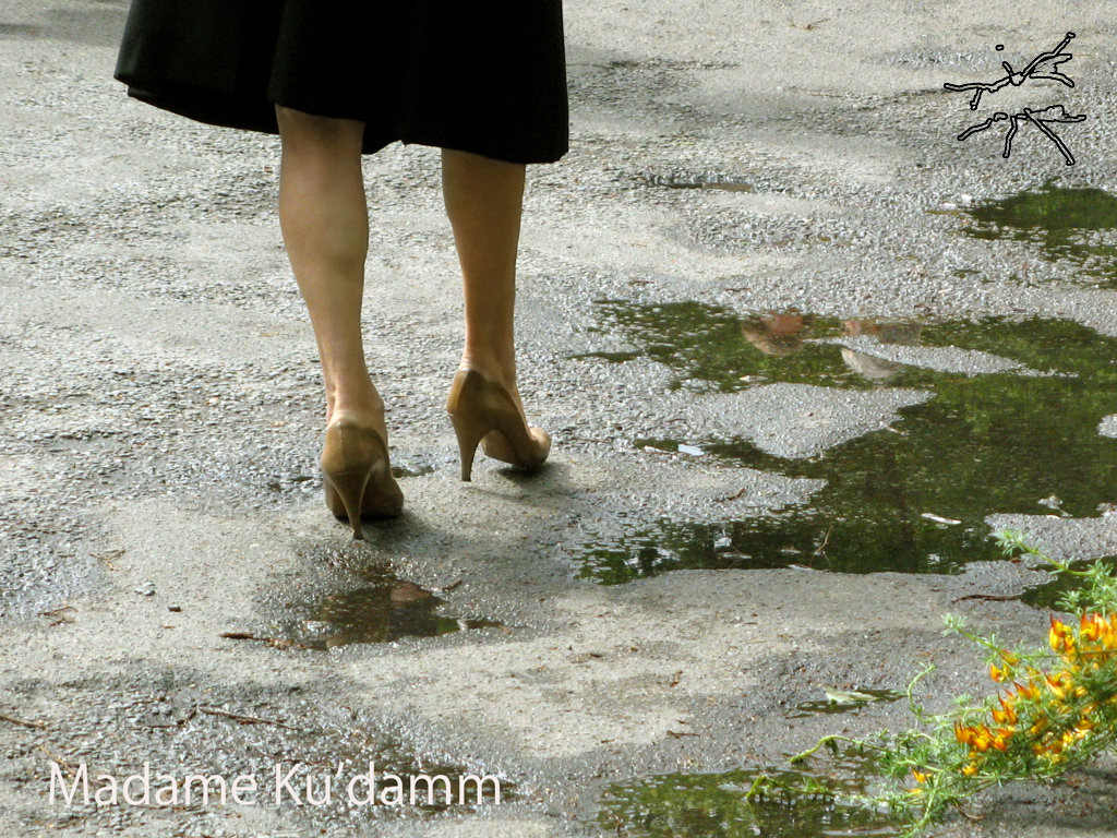 Eine Frau in beigefarbenen Pumps geht auf nassem Asphalt, umgeben von Pfützen. EN: A woman in beige high heels walks on wet asphalt, surrounded by puddles.