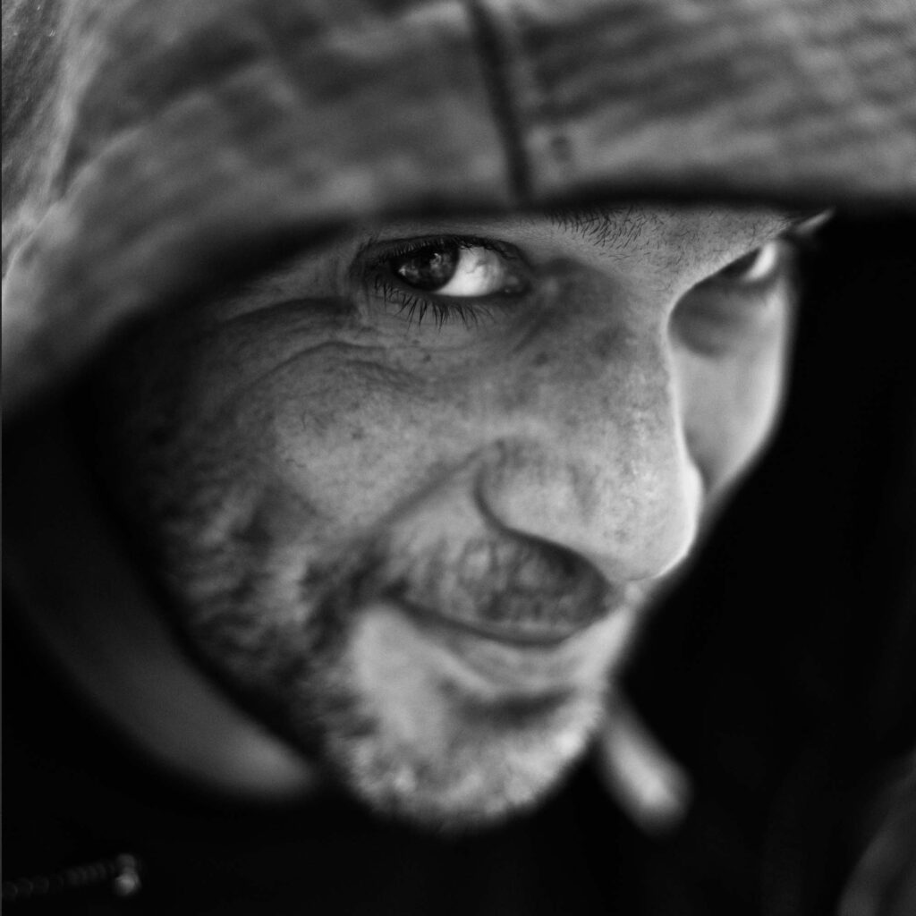 Alternativtext (Deutsch): Ein Blick aus der Tiefe der Kapuze – spielerisch, lauernd, vertraut. Das Licht bricht sich in den Augen, ein Schimmer zwischen Nähe und Distanz. Die Mundwinkel verraten mehr, als sie verbergen, ein Lächeln, das fast zum Gespräch wird. Die Fotografin Eliza fängt nicht nur einen Moment ein, sondern eine Verbindung – ein stilles Band zwischen Fotograf und Motiv. Alternative Text (English): A gaze from beneath the hood—playful, watchful, familiar. Light reflects in the eyes, a shimmer between closeness and distance. The corners of the mouth reveal more than they hide, a smile almost turning into a conversation. Photographer Eliza captures more than just a moment; she captures a connection—a silent bond between photographer and subject.