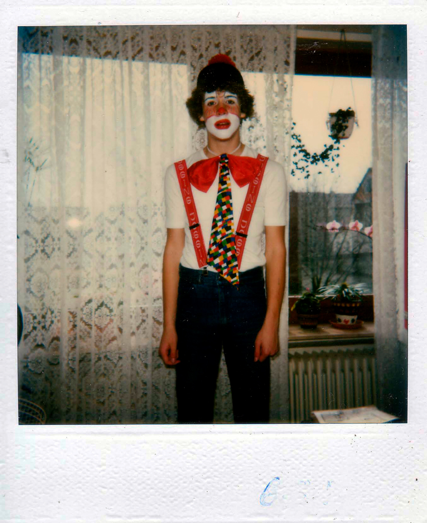 Deutsch: Ein Polaroid zeigt eine als Clown verkleidete Person mit rot-weißer Schminke, einer übergroßen Schleife und Hosenträgern. Die Pose wirkt steif, der Blick unsicher – eine Momentaufnahme von Verkleidung und Unbehagen. Der Raum dahinter bleibt vertraut, doch die Veränderung fühlt sich fremd an. English: A Polaroid captures a person dressed as a clown, with red and white face paint, an oversized bow, and suspenders. The posture seems stiff, the gaze uncertain – a snapshot of disguise and discomfort. The room behind remains familiar, yet the transformation feels strange.