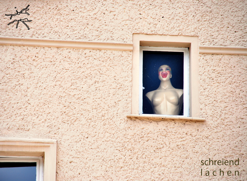 Mannequin-Oberkörper mit roten Lippen schaut lachend aus einem Fenster in einer beigen, strukturierten Wand. Mannequin torso with red lips looking out laughing from a window in a beige, textured wall.