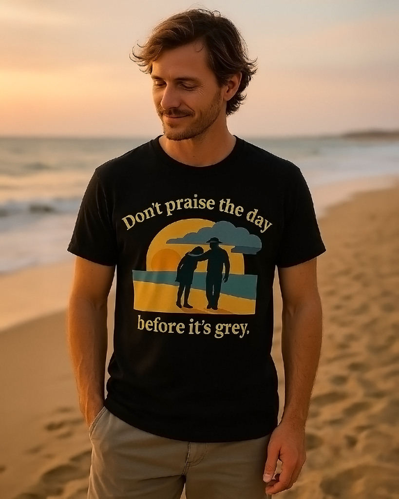 (DE) Person am Strand trägt ein T-Shirt mit der Aufschrift „Don’t praise the day before it’s grey“ und einem stilisierten Sonnenuntergangsmotiv. (EN) Person on the beach wearing a T-shirt with the text “Don’t praise the day before it’s grey” and a stylized sunset design.