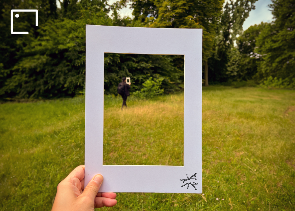 Eine Hand hält einen weißen Papierrahmen in die Wiese – darin sieht man eine Person mit Rucksack, die ebenfalls einen identischen Rahmen hält. Im Vordergrund das Fliegensymbol unten rechts im Rahmen.